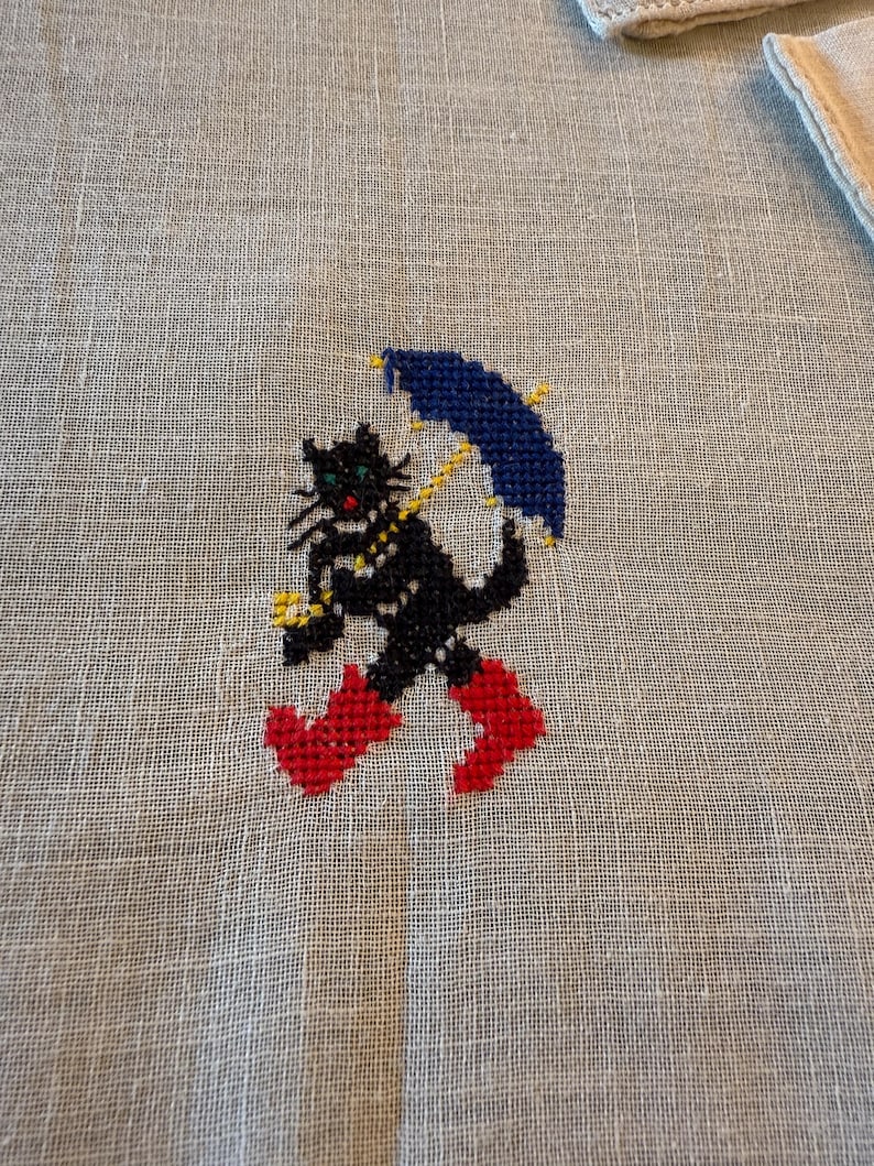 May include: A cross-stitched design of a black cat wearing red boots and holding a blue umbrella with yellow accents. The cat has green eyes and a yellow collar. The design is on a light beige linen fabric.