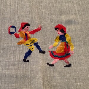 May include: Cross-stitch embroidery featuring two figures in traditional folk attire. One figure holds a tambourine. The figures are dressed in red, yellow, blue, and black. The embroidery is on a light gray fabric.