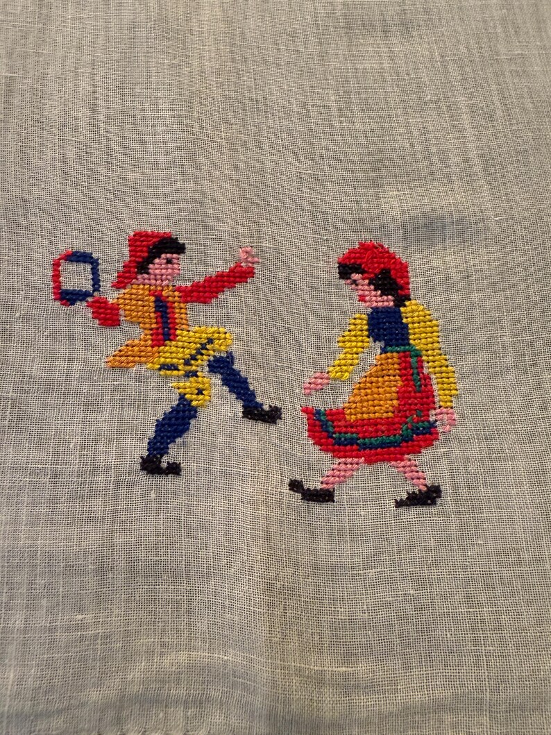 May include: Cross-stitch embroidery featuring two figures in traditional folk attire. One figure holds a tambourine. The figures are dressed in red, yellow, blue, and black. The embroidery is on a light gray fabric.