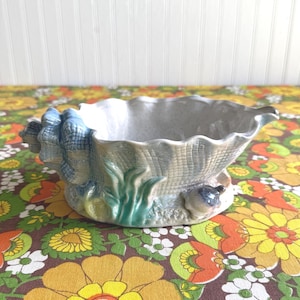 May include: A ceramic seashell-shaped planter with a scalloped rim. The planter is primarily gray with blue and green accents, including a detailed shell design and aquatic elements. It sits on a colorful floral patterned surface.