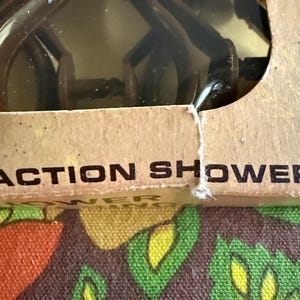 May include: A brown plastic shower head is visible through a clear plastic window in a cardboard box. The box has the words "ACTION SHOWER" printed on it. The background is a colorful floral pattern.