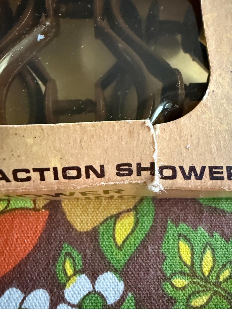 May include: A brown plastic shower head is visible through a clear plastic window in a cardboard box. The box has the words "ACTION SHOWER" printed on it. The background is a colorful floral pattern.