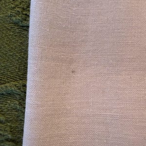 May include: Close-up of a folded, light-colored linen fabric. The fabric has a fine, textured weave and a subtle, neutral tone. The edge of the fabric is visible, revealing its folded construction. A green, textured surface is visible on the left side.