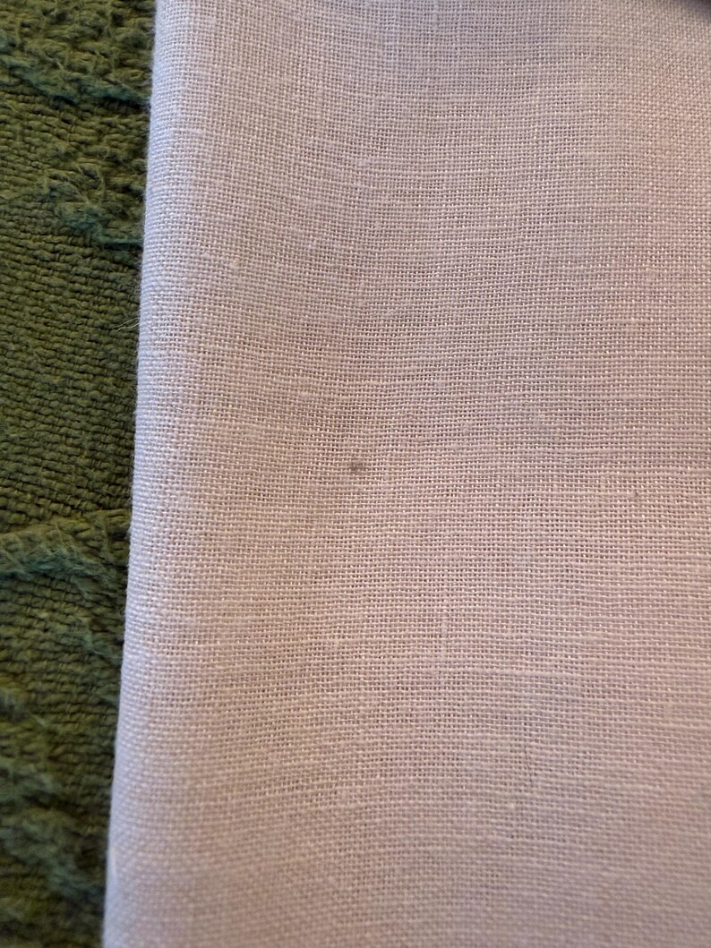 May include: Close-up of a folded, light-colored linen fabric. The fabric has a fine, textured weave and a subtle, neutral tone. The edge of the fabric is visible, revealing its folded construction. A green, textured surface is visible on the left side.