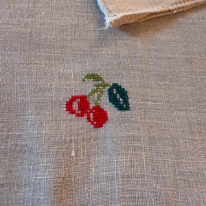 May include: A white linen handkerchief with a cross-stitched design of two red cherries with green leaves and stems. A corner of a second handkerchief with a floral design is visible in the background.