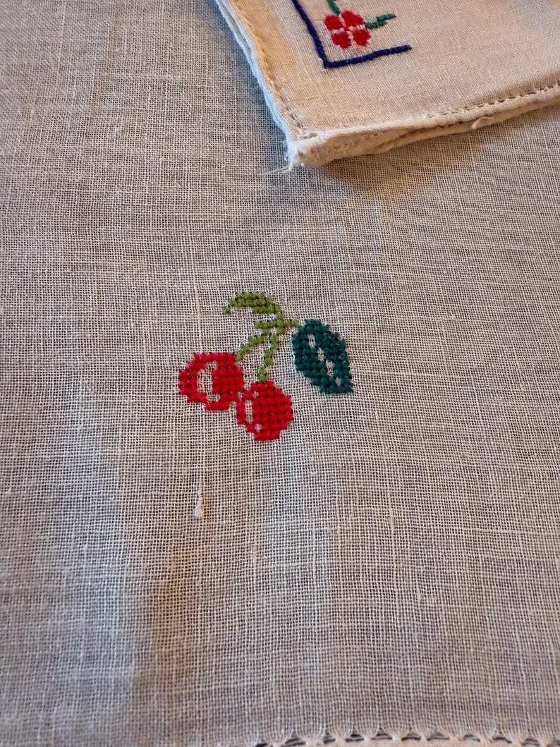 May include: A white linen handkerchief with a cross-stitched design of two red cherries with green leaves and stems. A corner of a second handkerchief with a floral design is visible in the background.