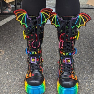 Può includere: Stivali in pelle nera con fibbie e lacci arcobaleno. Gli stivali hanno ali di pipistrello attaccate alla parte superiore.