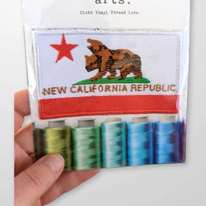 May include: A white patch with a red star and a brown bear with the text "NEW CALIFORNIA REPUBLIC" in red and green. The patch is packaged with five spools of thread in shades of green and blue.