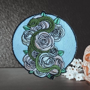 Puede incluir: Parche bordado con una serpiente verde enrollada alrededor de rosas blancas con hojas verdes, sobre un fondo circular azul claro. El parche tiene un borde negro, ideal para ropa o accesorios.
