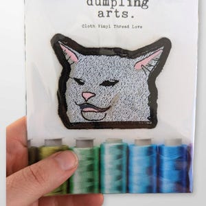 Könnte beinhalten: Ein grauer gestickter Bügel-Patch mit dem Meme "Frau schreit Katze an". Der Patch befindet sich auf einer weißen Karte mit dem Text "dumping arts. Cloth Vinyl Thread Love".