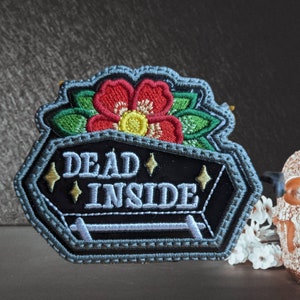 May include: Embroidered patch featuring a black coffin design with the words "DEAD INSIDE" in white. A red flower with green leaves and yellow center sits above the coffin. The patch has a gray border and gold star accents.
