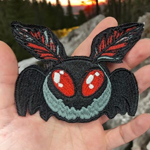May include: Black and red embroidered patch of a moth with large, menacing eyes. The moth has a jagged, toothy mouth.