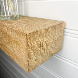 May include: A close-up of a light brown wooden shelf with a rough, textured surface. The shelf is mounted to a white wall with white wainscoting.