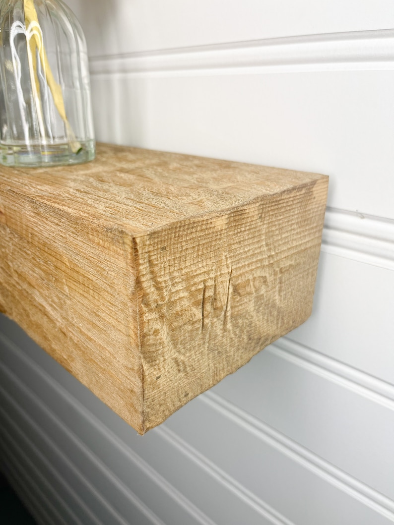 May include: A close-up of a light brown wooden shelf with a rough, textured surface. The shelf is mounted to a white wall with white wainscoting.