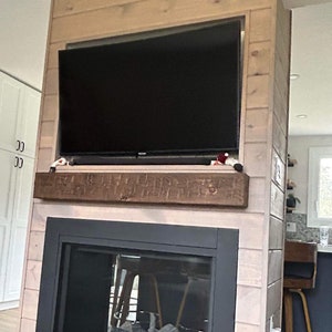 May include: A modern fireplace with a black metal frame and a glass front. The fireplace is surrounded by light wood panelling and has a wood mantel above it. There is a television mounted above the mantel.