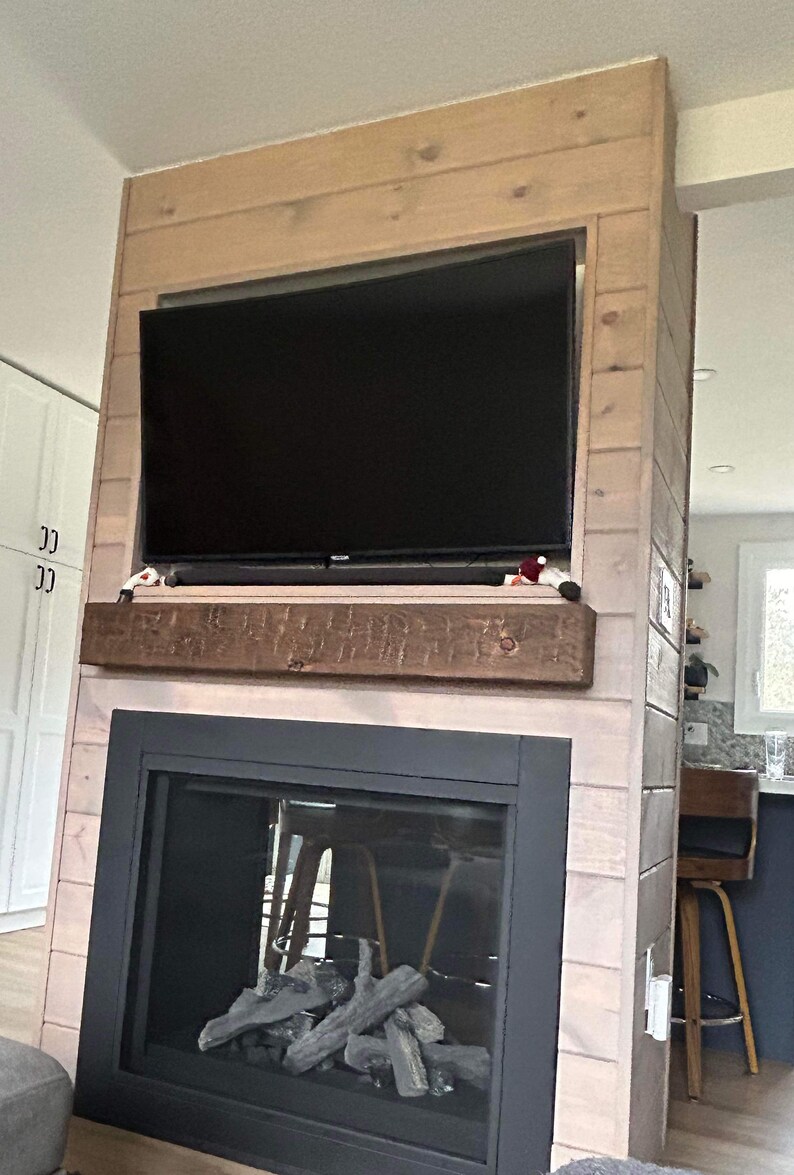 May include: A modern fireplace with a black metal frame and a glass front. The fireplace is surrounded by light wood panelling and has a wood mantel above it. There is a television mounted above the mantel.