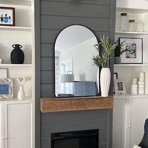 May include: A grey shiplap wall with a black arched mirror above a fireplace. The fireplace has a black surround and a black electric fire insert. A wooden mantel sits above the fireplace. There are white shelves on either side of the fireplace with various decorative items.
