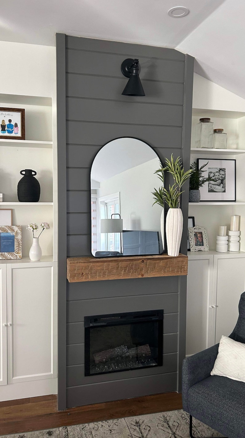 May include: A grey shiplap wall with a black arched mirror above a fireplace. The fireplace has a black surround and a black electric fire insert. A wooden mantel sits above the fireplace. There are white shelves on either side of the fireplace with various decorative items.