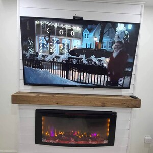 May include: A white fireplace mantel with a rustic wood shelf and an electric fireplace insert. The fireplace is decorated with a TV mounted above it.