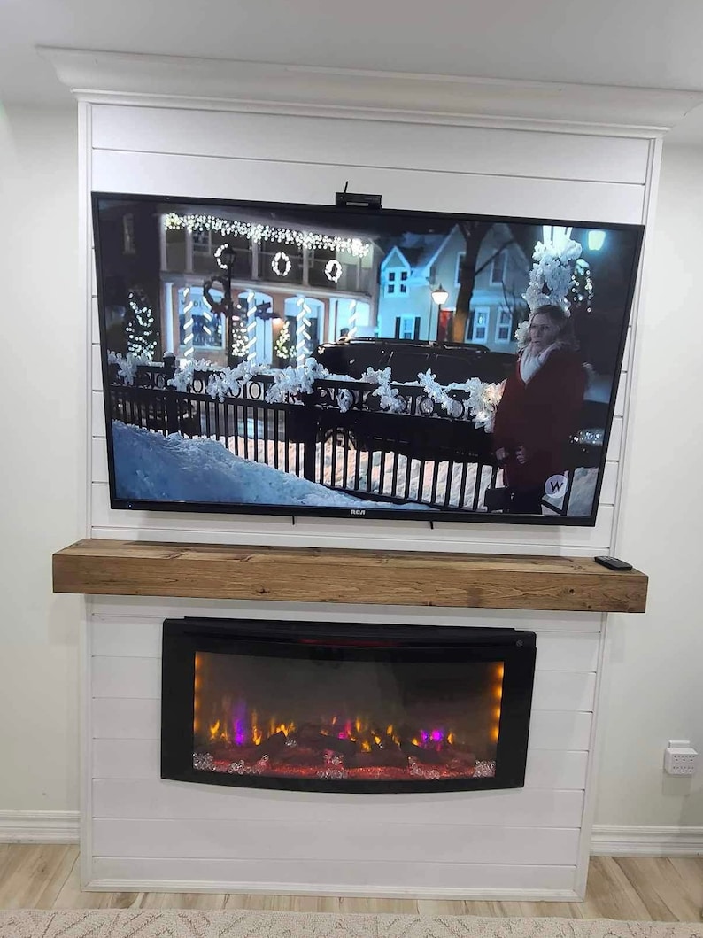 May include: A white fireplace mantel with a rustic wood shelf and an electric fireplace insert. The fireplace is decorated with a TV mounted above it.