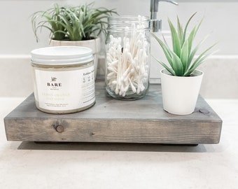 May include: A gray wooden tray with a white jar of lemon and ginger sugar scrub, a glass jar of cotton swabs, and a small potted succulent plant.