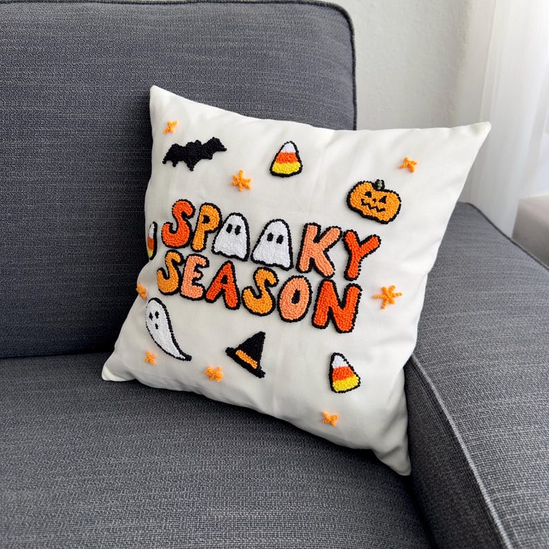 May include: White throw pillow with the words "Spooky Season" in orange and black puff letters. The pillow is decorated with ghosts, bats, candy corn, and a pumpkin.