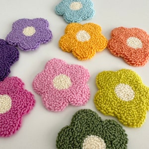 May include: A set of eight colorful flower-shaped coasters with white centers. The coasters are made of yarn and have a fluffy texture.