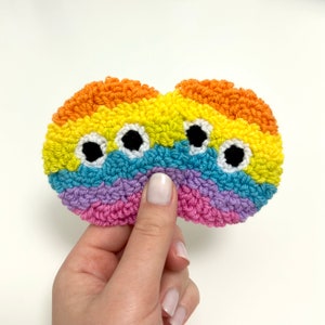 May include: A rainbow-colored, hand-stitched, felt eye mask with three black eyes. The mask is made of yarn and is held in a hand.