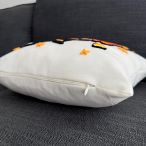 May include: White pillow with a zipper closure. The pillow has an orange and black embroidered design with small orange stars.
