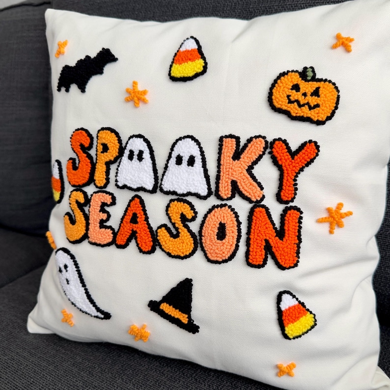 May include: White pillow with the words "Spooky Season" in orange, white, and black puff yarn. The pillow is decorated with ghosts, bats, candy corn, and pumpkins.
