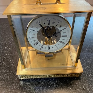 May include: A gold-toned, rectangular clock with a handle. The clock face is white with black Roman numerals and delicate black hands. The clock is encased in glass and sits on a gold base. The words "Seth Thomas" are printed on the clock.