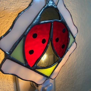May include: Stained glass night light shaped like a ladybug. The ladybug has a red body with black spots, surrounded by a white and yellow flower-like frame. The night light plugs into a standard wall outlet.