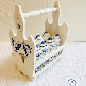 May include: A white wooden spice rack with a handle, holding six small ceramic spice jars. The jars and rack are decorated with blue floral patterns. The jars are labeled with spice names.