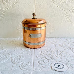 May include: A copper-colored canister with a lid and a small handle. The canister has a silver band around the middle and a small rectangular label that reads "KAFE". The canister is sitting on a white surface.