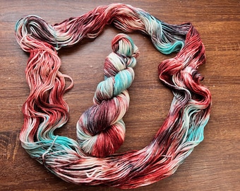 Puede incluir: Una madeja de hilo teñida a mano y un ovillo suelto a juego, ambos con una paleta de colores variados de rojo carmesí intenso, blanco crema suave, negro carbón oscuro y azul turquesa brillante, dispuestos en un patrón circular.