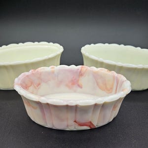 Op de afbeelding: Drie ovale keramische kommen met geschulpte randen. Eén kom is wit met roze en rode marmering, de andere twee zijn lichtgeel. De kommen staan op een zwart oppervlak.