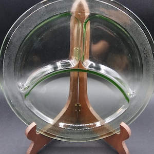 May include: A clear glass divided serving dish with a green accent line and a decorative dotted border. The dish is displayed on a wooden stand. The dish is likely vintage and suitable for serving appetizers or snacks.