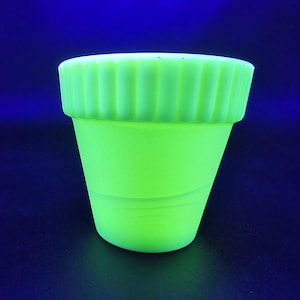 Puede incluir: Una maceta pequeña de color verde neón con un borde acanalado. La maceta tiene una forma ligeramente cónica y está hecha de un material sólido y opaco. El fondo es azul oscuro.