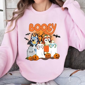 May include: A light pink crewneck sweatshirt featuring a graphic print of cartoon dogs in Halloween costumes. The text BOOEY is printed in orange above the characters, which include two ghosts, a vampire, and a pumpkin, surrounded by pumpkins.