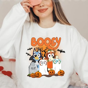 May include: A white crewneck sweatshirt featuring a graphic print with the text BOOEY in orange. The design includes two cartoon dogs dressed as a vampire and a pumpkin, two ghost figures, a tombstone marked RIP, bats, and pumpkins.