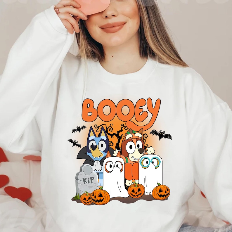 May include: A white crewneck sweatshirt featuring a graphic print with the text BOOEY in orange. The design includes two cartoon dogs dressed as a vampire and a pumpkin, two ghost figures, a tombstone marked RIP, bats, and pumpkins.