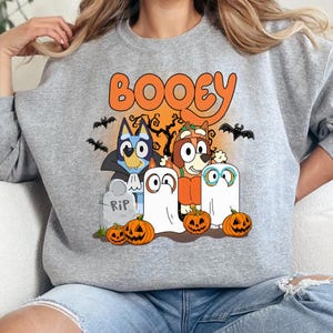 May include: A heather gray crewneck sweatshirt featuring a colorful graphic print with the text BOOEY. The design includes two cartoon dogs dressed in Halloween costumes, two ghost figures, three jack-o'-lanterns, bats, and a tombstone.