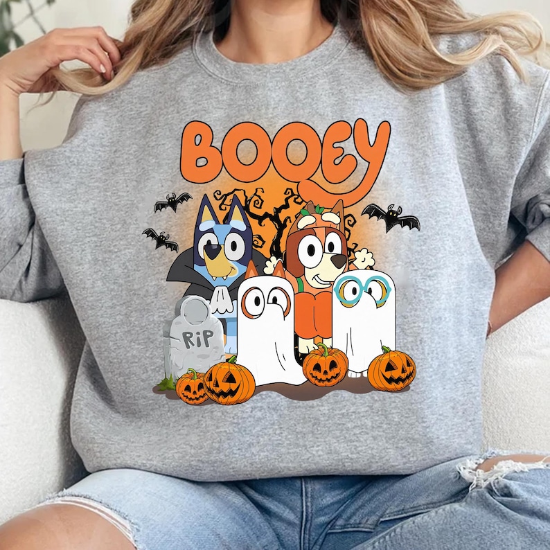 May include: A heather gray crewneck sweatshirt featuring a colorful graphic print with the text BOOEY. The design includes two cartoon dogs dressed in Halloween costumes, two ghost figures, three jack-o'-lanterns, bats, and a tombstone.
