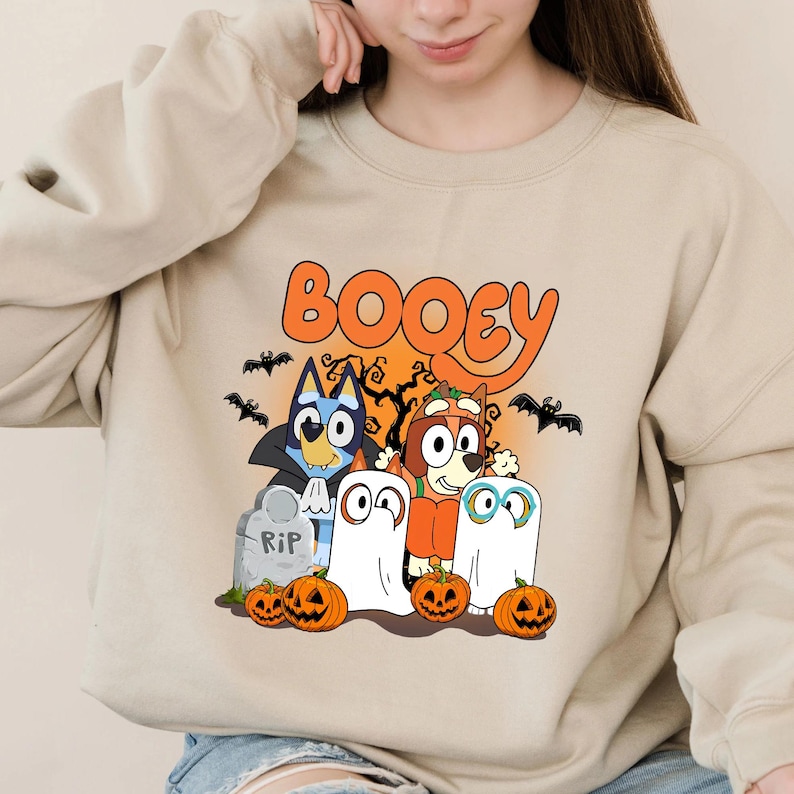 May include: A beige crewneck sweatshirt featuring a graphic print of two cartoon dogs dressed in Halloween costumes. The design includes the text BOOEY, a tombstone labeled RIP, orange pumpkins, and black bats against an orange moon background.