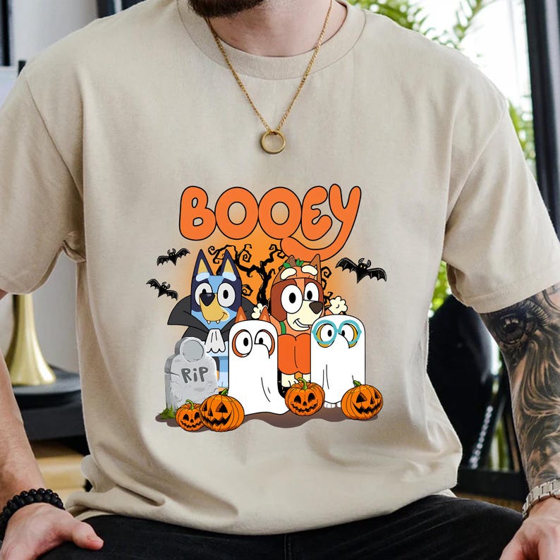 May include: A beige short-sleeve t-shirt featuring a graphic print with the text BOOEY in orange letters. The design includes two cartoon dogs dressed in Halloween costumes, two white ghost figures, a tombstone marked RIP, and orange pumpkins.
