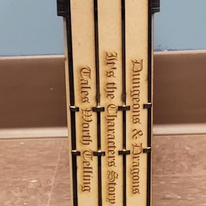 May include: A wooden Dungeons & Dragons dice tower with engraved text. The tower is rectangular with black accents and features the D&D logo. The text includes "Dungeons & Dragons", "It's the Character's Story", and "Tales Worth Telling".