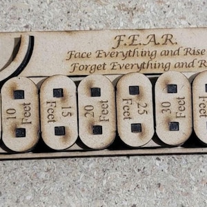 Puede incluir: Placa de madera con la inscripción F.E.A.R. (Face Everything and Rise Or Forget Everything and Run) y ocho marcadores de distancia extraíbles, de 1,5 a 12 metros. La placa tiene una parte superior curvada y es de madera clara.