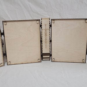 May include: A wooden box with four hinged panels, each with a light brown finish. The box is open, revealing a compartment with a sword design and a series of slots for dice.