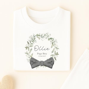 May include: White pyjama set with a t-shirt featuring a green floral wreath, a grey bow tie, and the name "Ollie" with "Page Boy" and a date. A wooden star and a sprig of leaves are also present.
