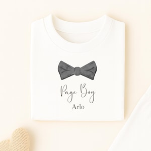 May include: A white baby's pajama set featuring a grey bow tie design and the words "Page Boy Arlo". The set comprises a long-sleeved top and trousers. A wooden star toy, a sprig of leaves, and a heart-shaped toy are also visible.